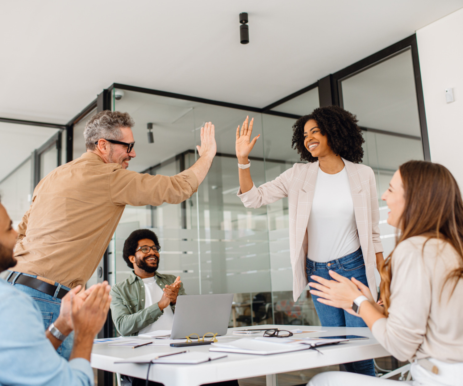A diverse team of professionals celebrates a workplace achievement with a high-five in a modern office. The moment captures motivation, cooperation, and a positive business culture focused on teamwork